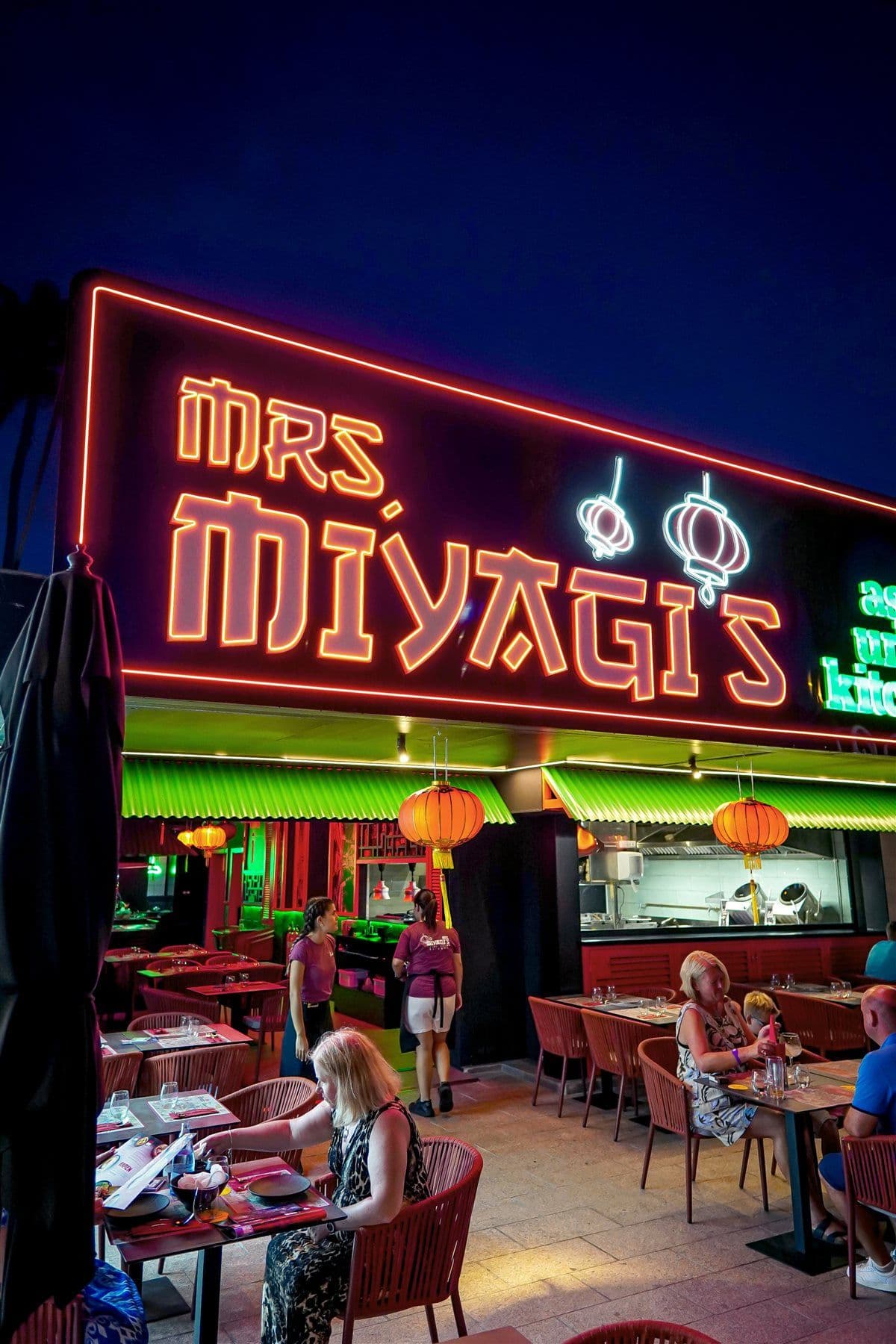 Fachada de Mrs Miyagi's de noche, con el rótulo de neón rojo y verde y los faroles sobre la terraza