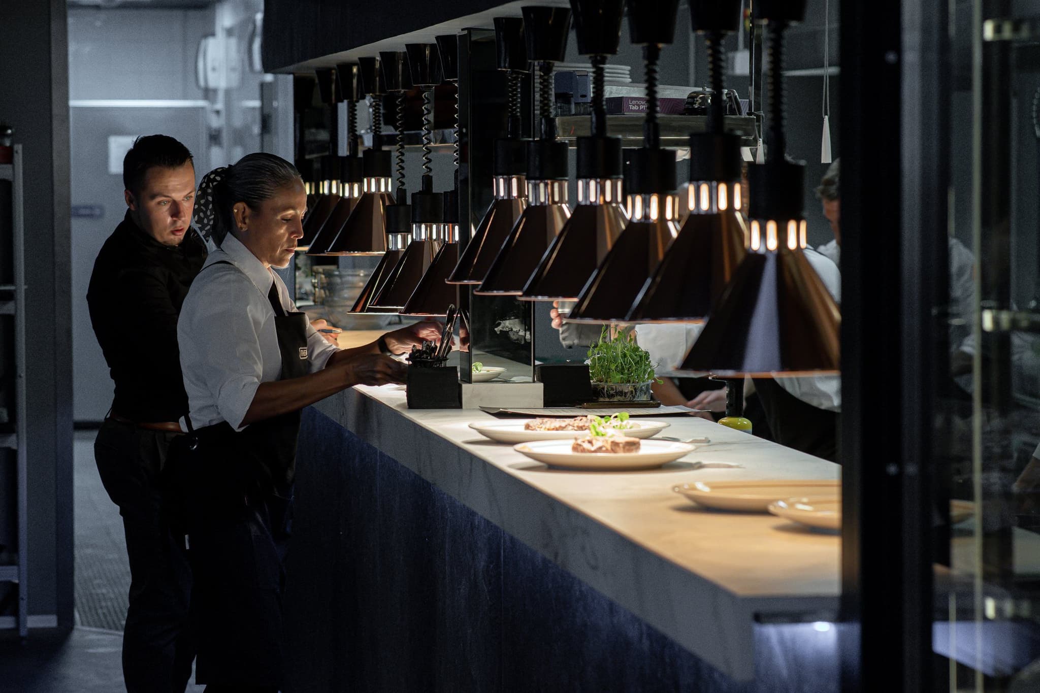 Pase de cocina de The Bank Steakhouse: fila de lámparas de calor sobre encimera de mármol, con dos personas del equipo emplatando durante el servicio