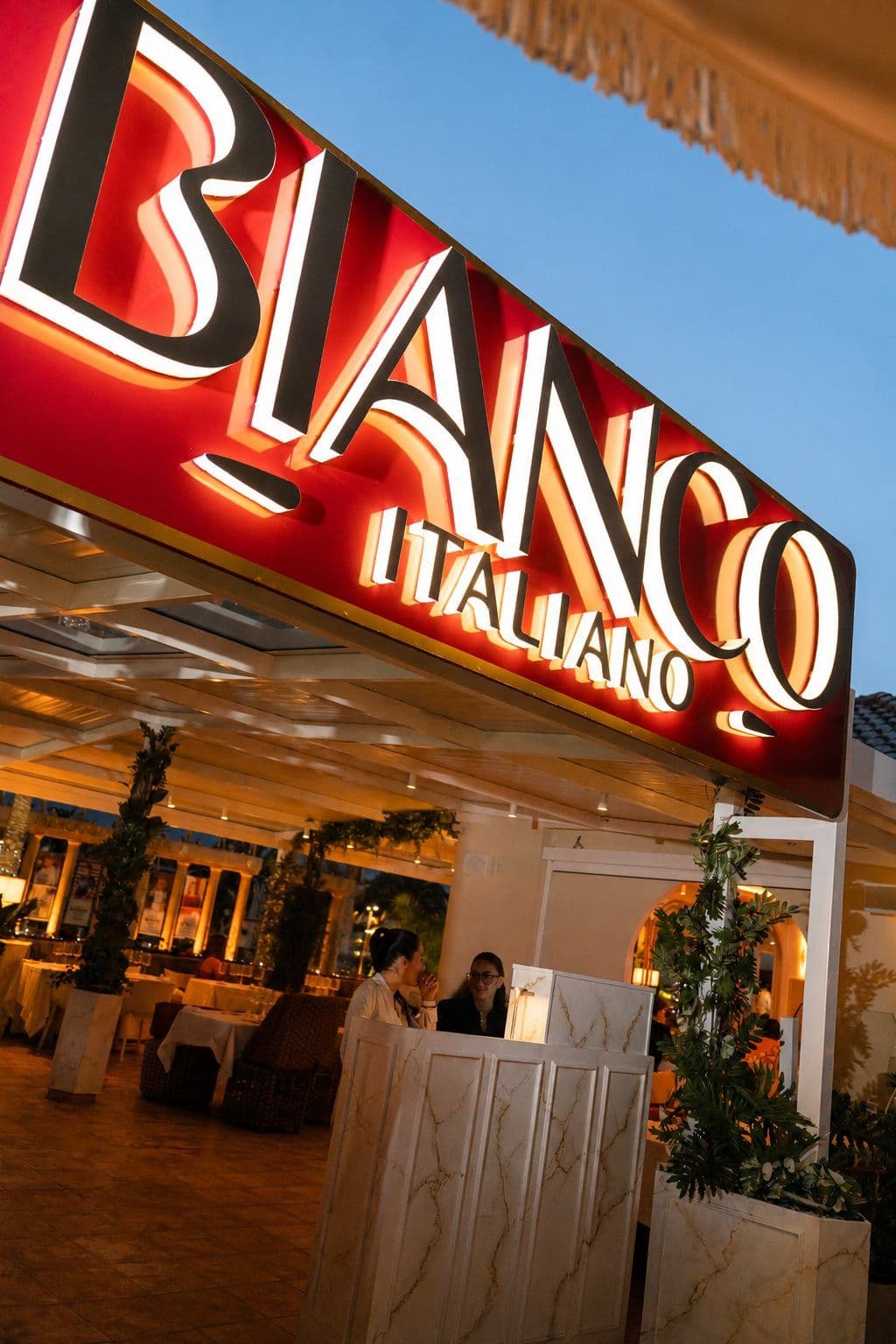 Fachada de Bianco Italiano iluminada de noche, con el rótulo en rojo y la sala al fondo