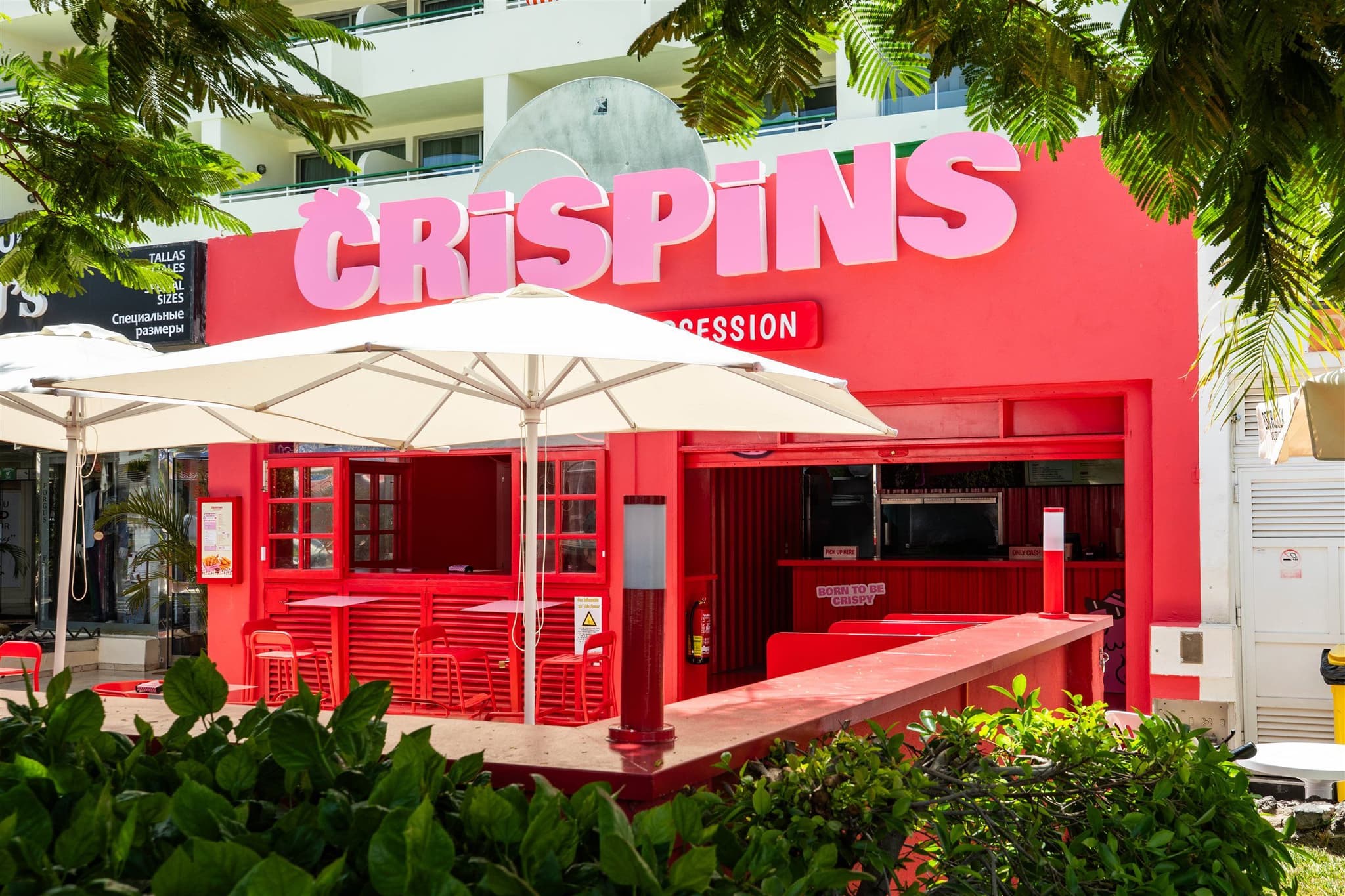 Fachada de Crispins con el rótulo en rosa sobre el frente rojo y la terraza cubierta por sombrillas