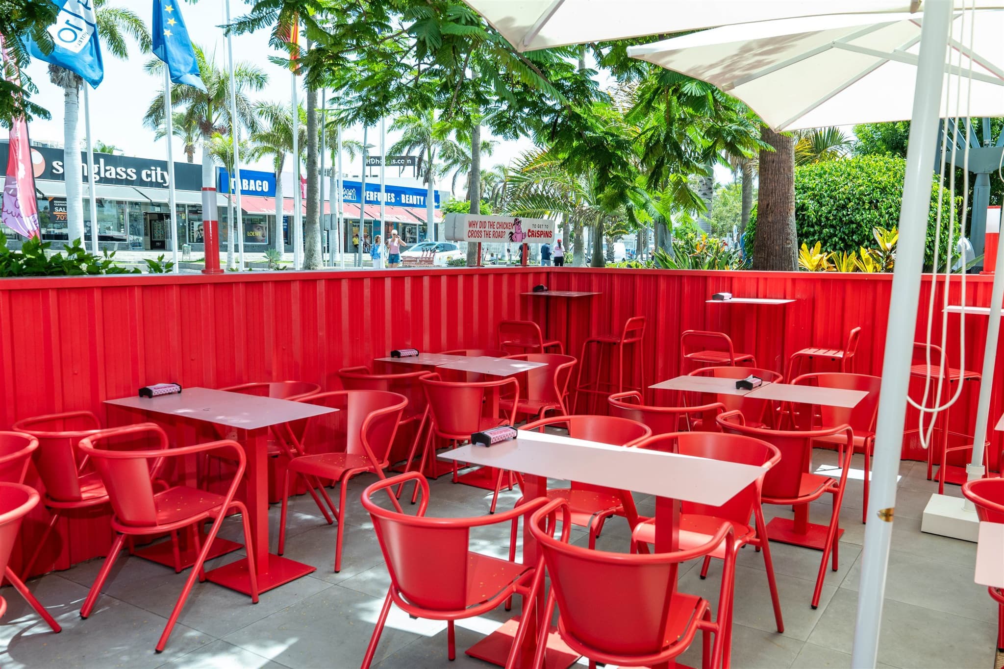 Terraza de Crispins con mesas y sillas rojas bajo las palmeras del paseo